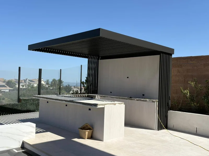 4K cantilever with 2-inch lattice in espresso, over a hillside outdoor kitchen with slat privacy screens and a glass view fence
